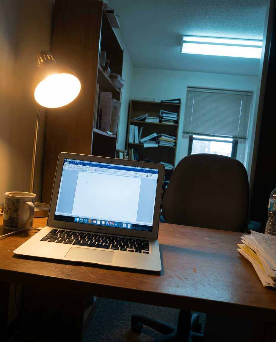 A laptop open on a desk with the cursor blinking on a mostly empty page, early afternoon, the chair pushed back and empty
